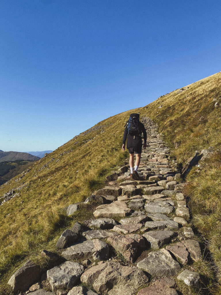 hiker walking up the stone path towards the summit of Ben Nevis, blue skies